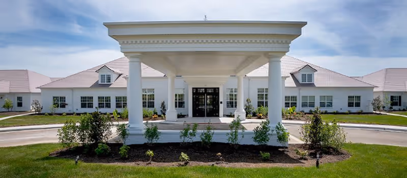 Front exterior view of Ashton Grove facility featuring a large white building with a covered entrance supported by four columns, surrounded by landscaped greenery and a circular driveway under a partly cloudy sky.