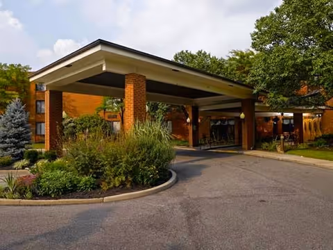 Covered entrance and driveway of a brick senior living facility with landscaped islands and trees.