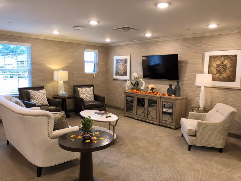A cozy living room with a beige sofa, two dark leather chairs, and a light-colored armchair arranged around two small coffee tables. The room features a flat-screen TV mounted on the wall above a wooden cabinet decorated with vases and flowers. Two framed artworks hang on the walls, and two table lamps provide warm lighting. A window with blinds lets in natural light.