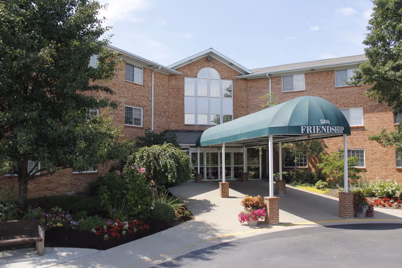 Exterior view of Friendship Towers, a three-story brick building with multiple windows. The entrance features a green canopy with the number 580 and the word 'FRIENDSHIP' on it. There are landscaped flower beds and trees around the entrance area.