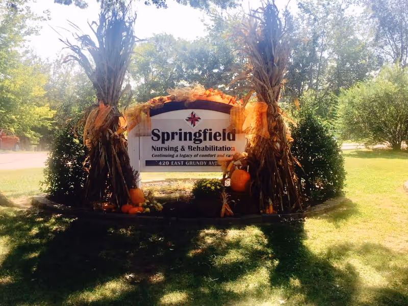 Outdoor sign for Springfield Nursing & Rehabilitation Center decorated with autumn-themed decorations including cornstalks, pumpkins, and orange ribbons, surrounded by greenery and trees in a sunny setting.