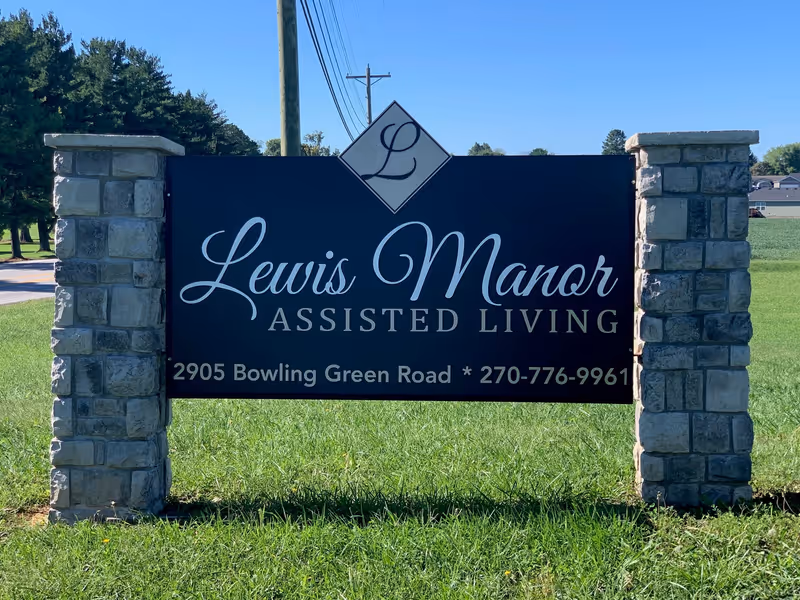 Outdoor sign for Lewis Manor Assisted Living mounted between two stone pillars on a grassy area with trees and a clear blue sky in the background.