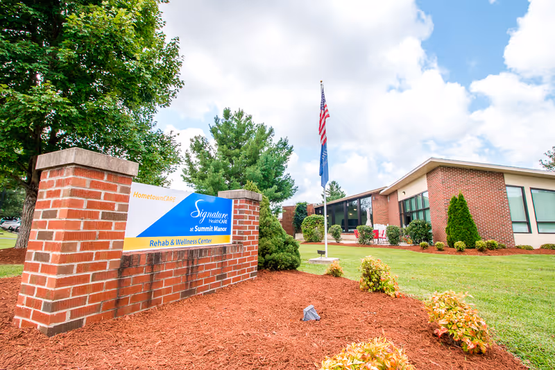 Brick entrance sign and landscaped front lawn of Signature HealthCARE at Summit Manor Rehab & Wellness Center with the facility building and flagpoles in the background.