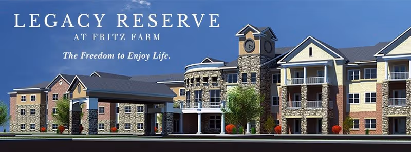 Exterior view of Legacy Reserve at Fritz Farm, a multi-story senior living facility with a clock tower feature, balconies, and a mix of stone and siding on the facade under a clear blue sky.