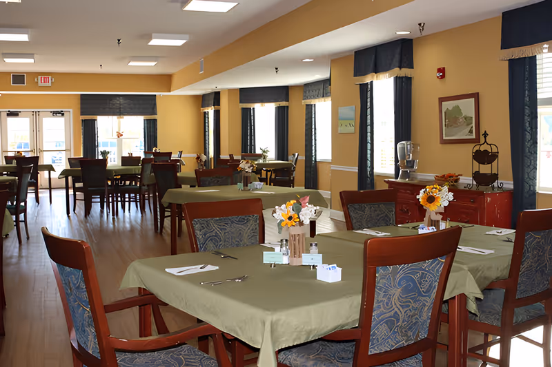 Bright communal dining room with tables covered in green tablecloths, wooden chairs, and floral centerpieces.