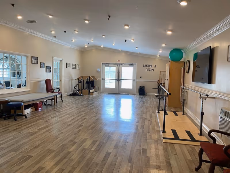 Wide, well-lit rehabilitation room with parallel bars, exercise equipment, and ample open floor space.