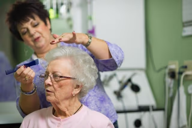 An elderly woman with white hair and glasses is seated while a hairdresser combs and styles her hair in a salon setting with green walls and hairdressing equipment in the background.