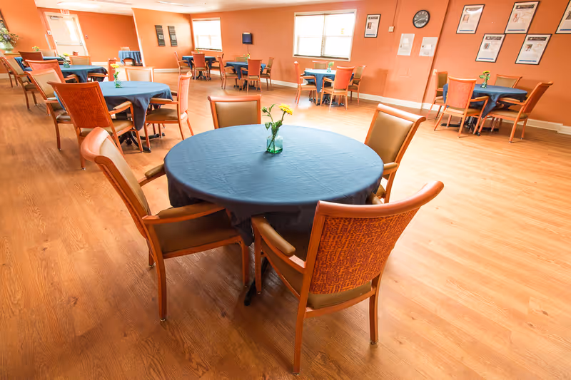 A spacious dining room with several round tables covered with blue tablecloths, each surrounded by wooden chairs with orange and brown upholstery. Small flower vases are placed on each table. The room has wooden flooring, peach-colored walls, windows letting in natural light, and framed posters on the walls.