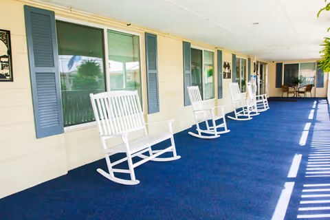 A covered outdoor porch area with several white wooden rocking chairs lined up along the wall of a building with windows and blue shutters. The floor is covered with a blue carpet, and there is a small table with chairs at the far end of the porch.