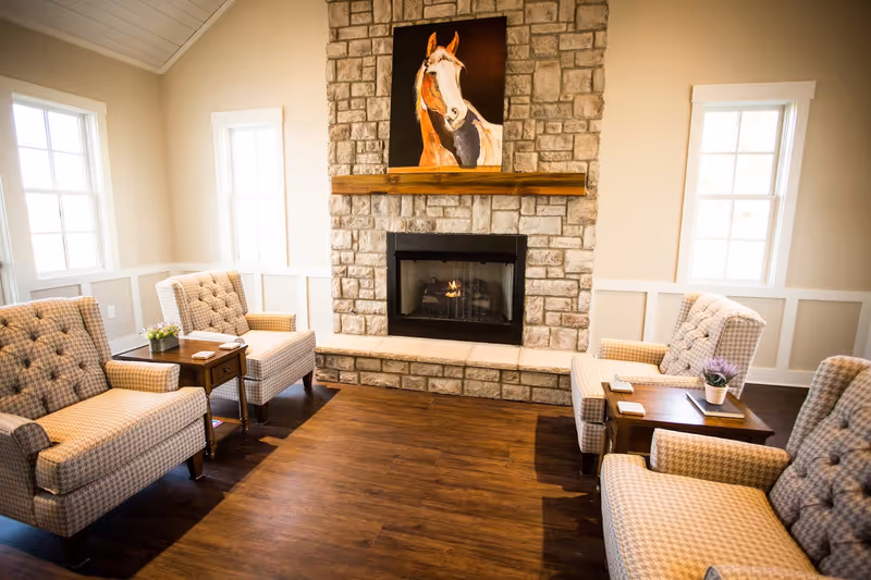 A cozy living room with four beige patterned armchairs arranged around a stone fireplace. The fireplace has a wooden mantel with a painting of a horse above it. There are two windows on either side of the fireplace letting in natural light. Small wooden side tables with plants and coasters are placed between the chairs on a wooden floor.