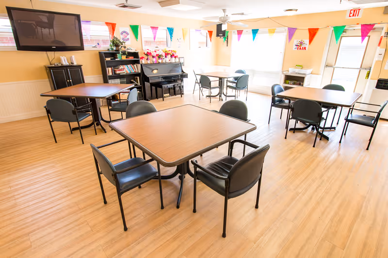A bright and clean common room with multiple square tables and black chairs arranged around them. The room has light wood flooring, a piano against the wall with flowers on top, a bookshelf with books, a large flat-screen TV mounted on the wall, and colorful triangular pennant banners hanging near the ceiling. Large windows allow natural light to fill the space.
