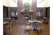 Community dining area with wooden tables and upholstered chairs facing a brick fireplace adorned with decorative items.