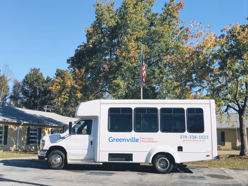 A white Greenville Nursing & Rehabilitation shuttle bus parked outside a facility with trees and an American flag in the background.