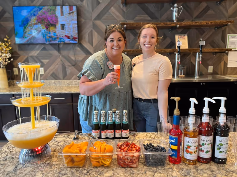Two women smiling behind a counter with a drink fountain, bottles of sparkling wine, containers of sliced fruit, and syrup bottles. A television displaying a scenic image is mounted on the wall behind them.