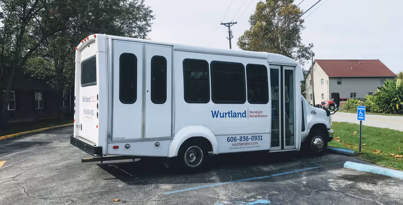 A white shuttle bus parked in a handicapped parking space outside, with the text 'Wurtland Nursing & Rehabilitation' and contact information on its side. Trees and buildings are visible in the background.
