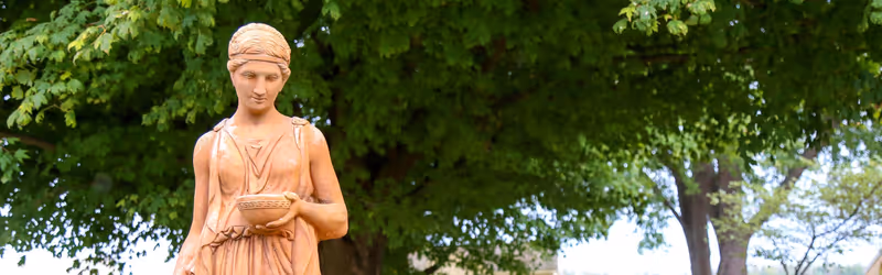 A terracotta-colored statue of a woman holding a bowl stands in a leafy outdoor area.