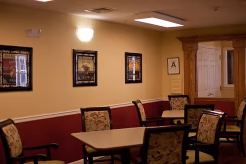 Interior view of a common area with a rectangular table surrounded by six upholstered chairs with floral patterns. The walls are painted beige and maroon with three framed pictures hanging on the wall. There is a wall-mounted light fixture and a wooden doorway leading to another room.