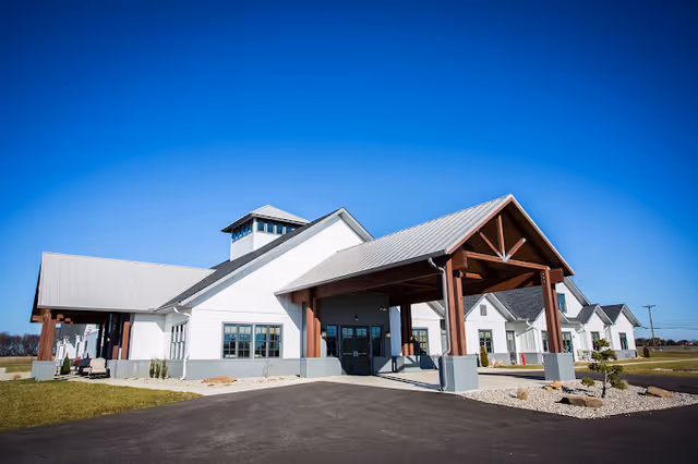 Exterior view of a modern senior living facility building with white walls, a gray metal roof, and wooden beams supporting a covered entrance. The building is surrounded by a paved driveway, landscaped areas with rocks and small plants, and a clear blue sky overhead.
