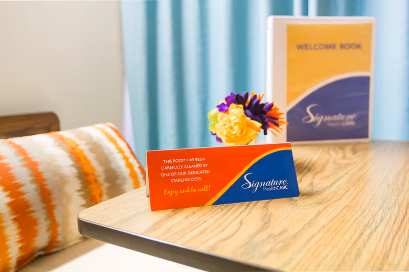 A wooden table in a care facility displays a Signature HealthCARE cleaned-room card, a small flower arrangement, and a welcome book with a patterned pillow and curtains in the background.
