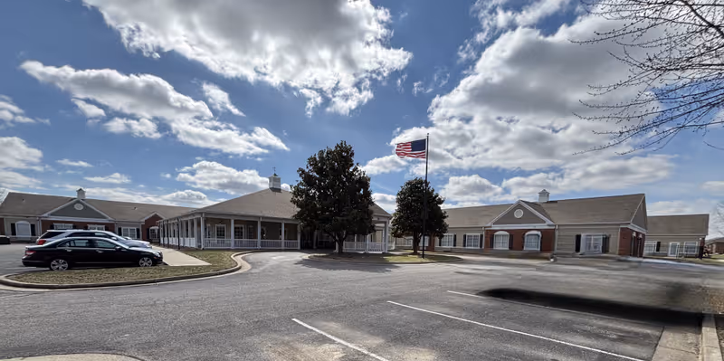 Exterior view of a single-story senior living facility building with a covered porch, several windows, and an American flag on a flagpole in front. There are a few parked cars and a partly cloudy sky overhead.