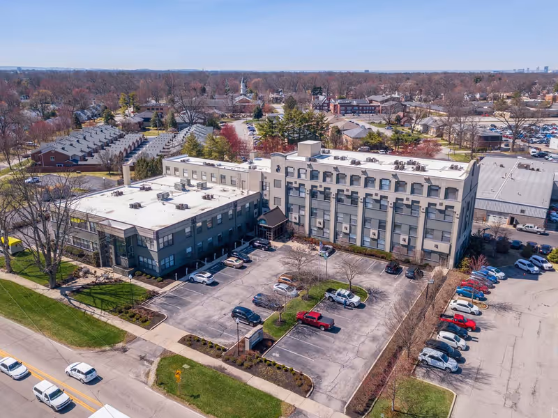 Aerial view of a multi-story building with a parking lot in front, surrounded by trees and residential houses in the background under a clear sky.