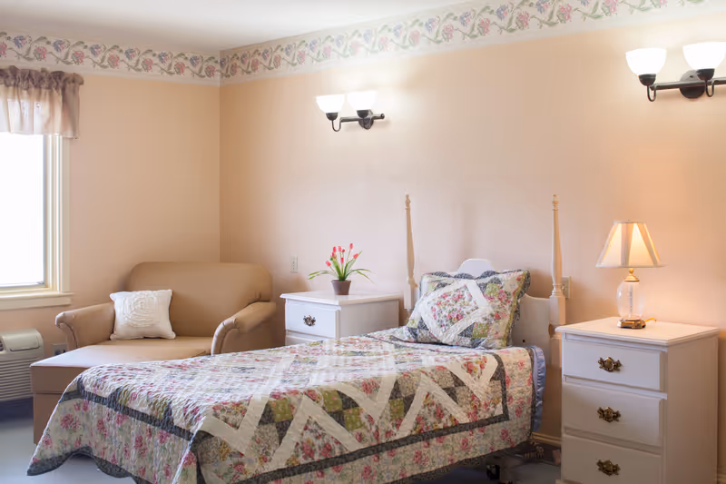 A cozy single bedroom with a floral quilted bed, an armchair by the window, nightstands with a lamp and flowers, and soft pastel walls.