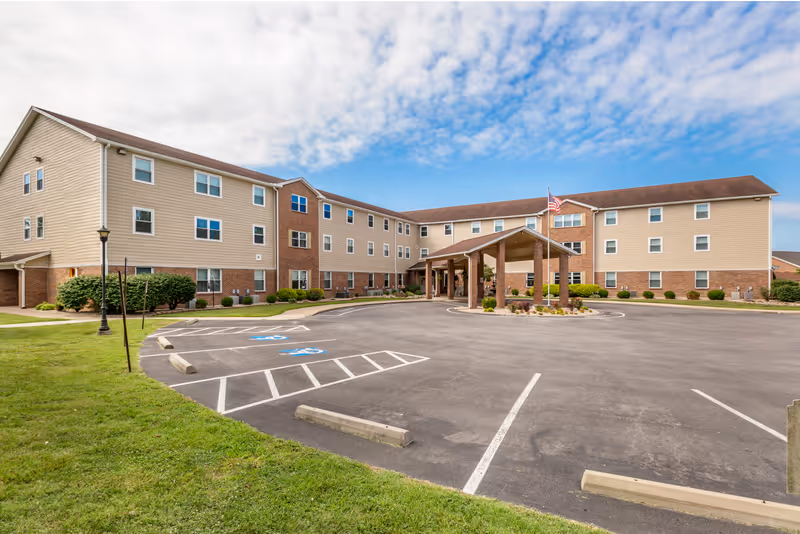 Exterior view of a three-story senior living facility building with beige siding and brick accents. The building has multiple windows and a covered entrance with an American flag on a flagpole. The foreground shows a parking lot with marked handicap parking spaces and green grass on the sides under a partly cloudy blue sky.