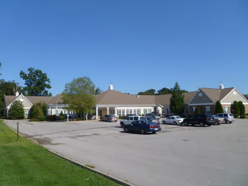 Exterior view of Fairview Place Senior Living facility showing a single-story building with a large parking lot in front. Several cars are parked, and there are trees and shrubs around the building under a clear blue sky.