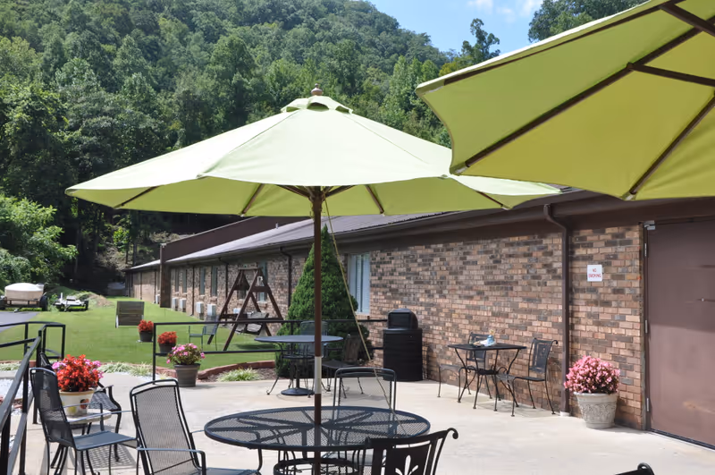 Outdoor patio area with black metal tables and chairs under large green umbrellas. There are potted flowers and a brick building wall in the background, with a grassy area and trees behind it.