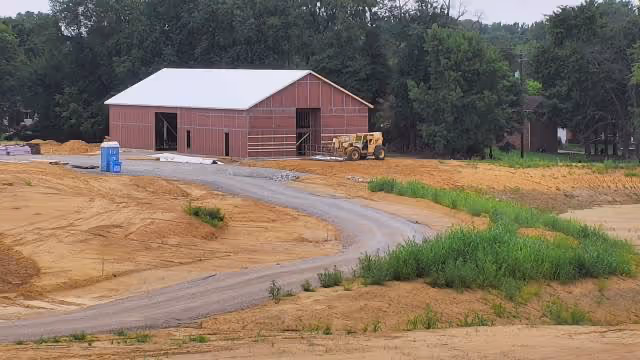 A construction site with a large red barn-like building with a white roof in the background. The ground is mostly dirt with some patches of green vegetation, and a gravel road curves toward the building. Trees are visible in the background.