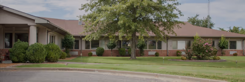 Single-story brick and siding building with a brown roof, surrounded by green grass, bushes, and trees under a partly cloudy sky.