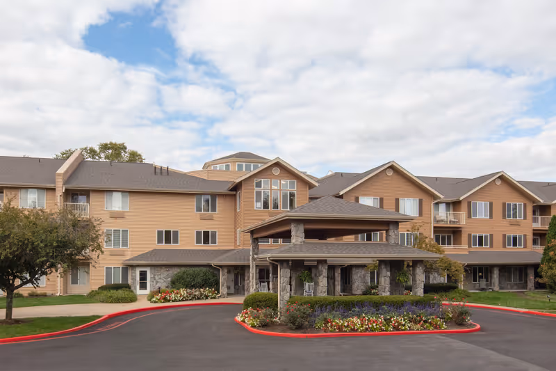 Covered front entrance and porte-cochere of a multi-story beige senior living building with landscaped flowerbeds and driveway.