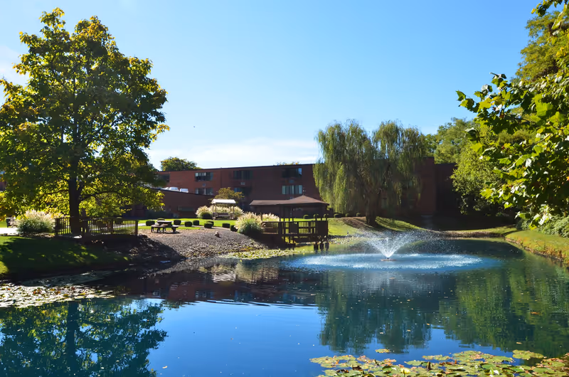 A peaceful outdoor scene at Brownsboro Park Retirement Community featuring a pond with a water fountain in the center, surrounded by lush green trees and bushes. There are wooden benches and picnic tables near the pond, with a brick building visible in the background under a clear blue sky.