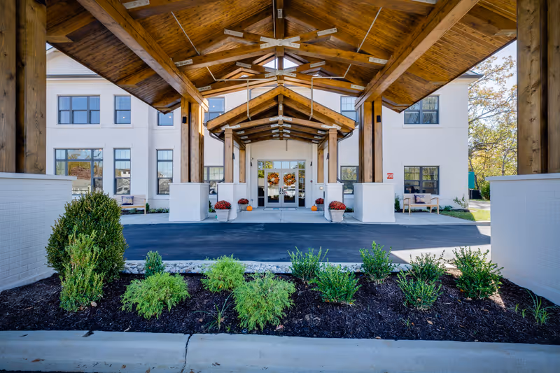 Entrance of a senior living facility with a wooden covered driveway, white brick exterior walls, large windows, and landscaped bushes in front. There are benches on either side of the entrance and autumn decorations on the doors.