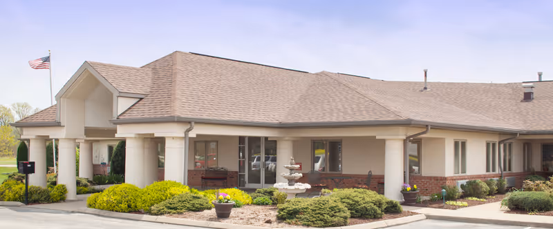 Exterior view of Heartland Villa Center, a single-story building with a beige and brick facade, a covered entrance supported by columns, landscaped bushes, a small fountain, and an American flag flying on a pole.