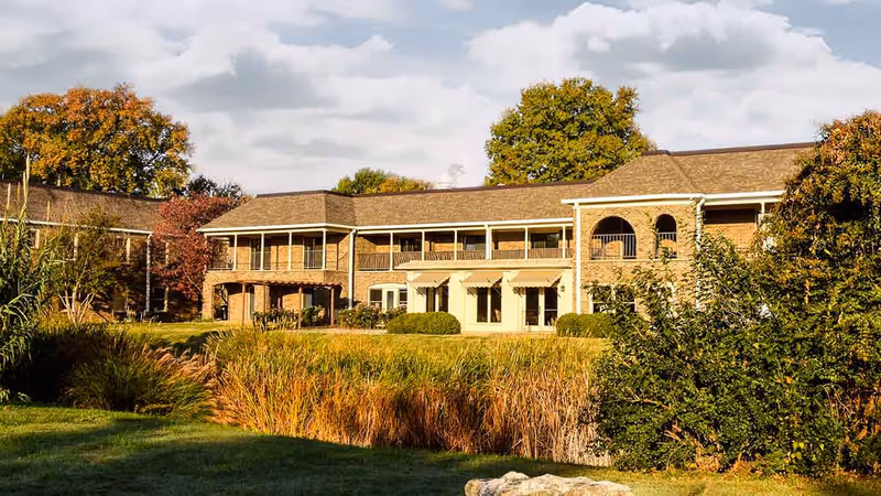 Exterior view of a two-story brick building with balconies and large windows, surrounded by trees and bushes with autumn foliage under a partly cloudy sky.