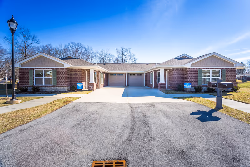Front exterior view of a single-story brick building with two garage doors in the center, green shutters on the windows, a street lamp on the left, and two mailboxes on the right. The sky is clear and blue, and there are leafless trees in the background.