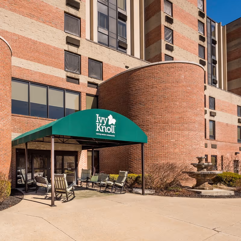Entrance of Ivy Knoll Senior Living Community with a green canopy displaying the facility's name, several cushioned chairs arranged under the canopy, a brick building facade, and a stone fountain to the right.