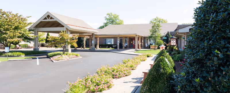 Front exterior view of Christian Health Center Hopkinsville showing a covered entrance driveway, landscaped flower beds, and bushes with a clear blue sky.
