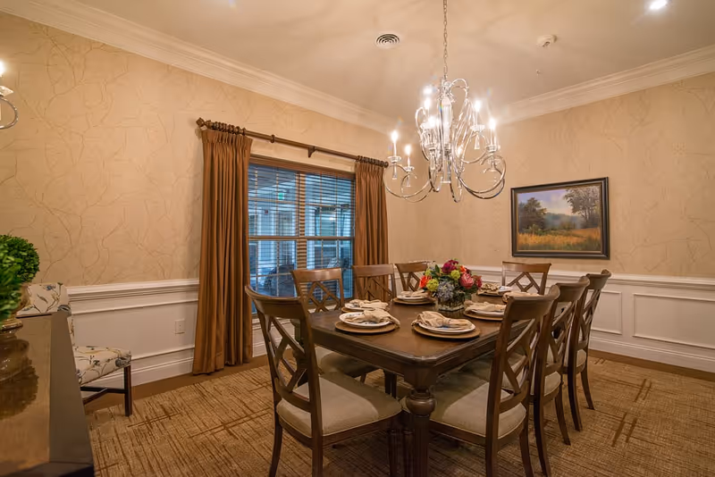 A warmly lit dining room with a wooden table set for eight people, featuring beige cushioned chairs, a floral centerpiece, and neatly arranged plates with napkins. The room has beige patterned wallpaper, white wainscoting, a window with brown curtains, a chandelier overhead, and a framed landscape painting on the wall.