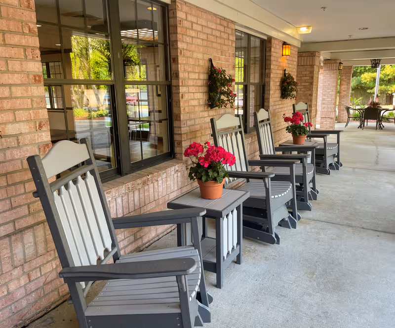 Covered outdoor patio area with a row of gray and white rocking chairs and small tables, each table holding a pot of bright pink flowers. The patio is adjacent to a brick building with large windows and hanging flower baskets. In the background, there is another seating area with a table and chairs surrounded by greenery.