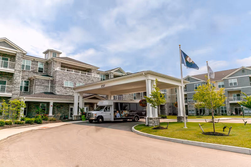 Exterior view of StoryPoint Union senior living facility showing a multi-story building with stone and siding facade, a covered entrance with a shuttle van parked underneath, landscaped greenery, and two flagpoles with flags waving in the breeze under a partly cloudy sky.