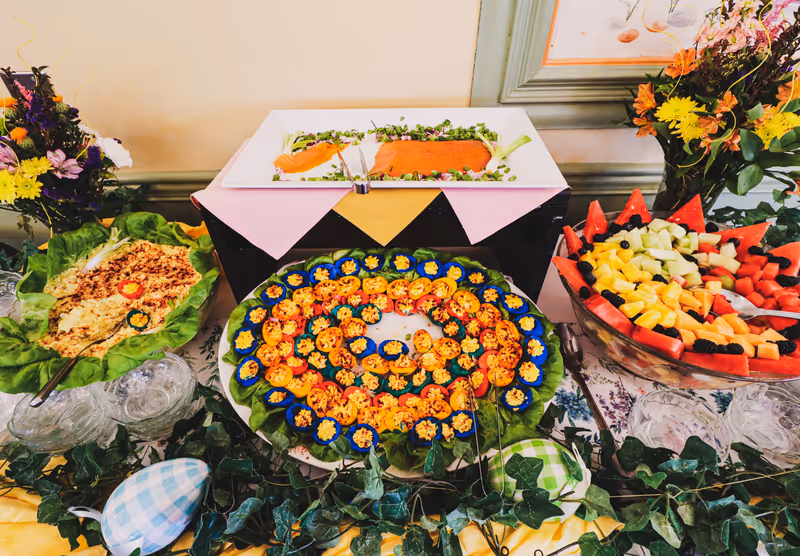 A buffet table with a variety of foods including a large bowl of mixed fruit, a platter of deviled eggs arranged in a spiral pattern, a dish of smoked salmon garnished with greens, and a bowl of salad. The table is decorated with green ivy and colorful flowers in vases.