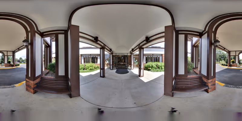 Covered entrance area of The Forum at Brookside senior living facility with brown wooden beams and benches on either side, concrete floor, and glass doors leading inside. Green bushes and trees are visible outside under a partly cloudy sky.