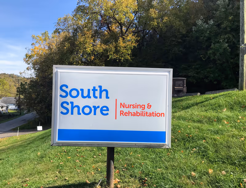 Outdoor sign for South Shore Nursing & Rehabilitation on a grassy area with trees and a road in the background under a clear blue sky.