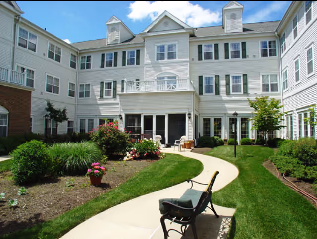 Outdoor courtyard area of a senior living facility with a curved concrete walkway, green grass, flower beds, bushes, and a bench with a cushion. The building surrounding the courtyard is white with multiple windows and a blue sky with some clouds above.