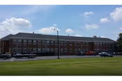 Brick nursing home exterior with parked cars and a grassy front lawn under blue sky.