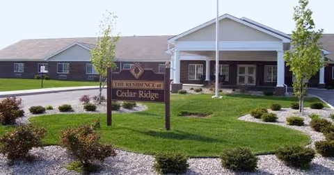 Exterior view of Cedar Ridge Health Campus showing a single-story building with a covered entrance, a flagpole, and landscaped grounds with grass, small bushes, and trees. A sign in front reads 'The Residence at Cedar Ridge.'