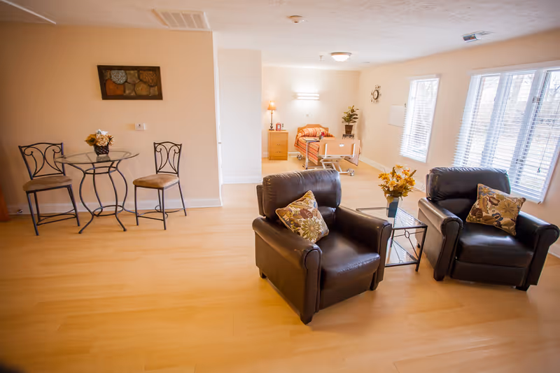 A bright and spacious room with light wood flooring featuring two black leather armchairs with floral cushions around a glass-top side table with a flower vase. In the background, there is a hospital-style bed with orange bedding, a wooden nightstand with a lamp, and a small table with a plant. To the left, there is a small round glass table with two metal chairs. Large windows with white blinds allow natural light to fill the room.