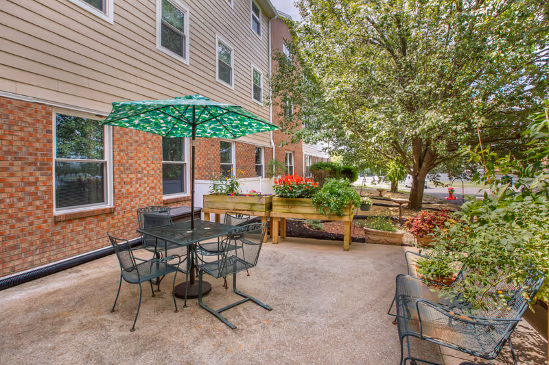 Outdoor patio area with a metal table and four chairs under a green patterned umbrella. Raised garden beds with colorful flowers and plants are nearby, along with additional potted plants and a large tree providing shade. The patio is adjacent to a brick and siding building with several windows.